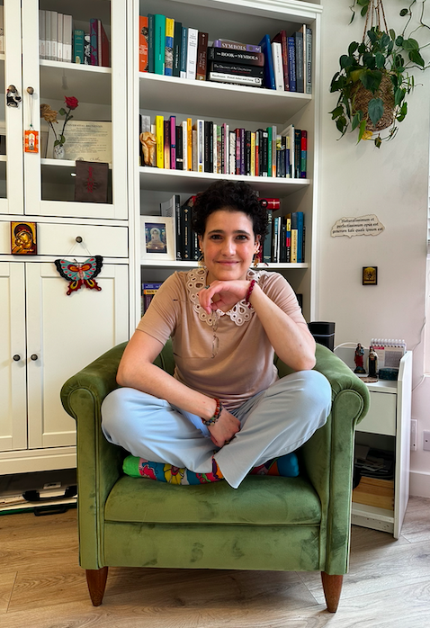 A person sits cross-legged on a green chair in a cosy, book-filled room.
