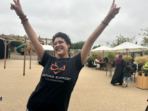 Person in a t-shirt celebrates with arms raised outdoors, smiling in a cheerful environment.