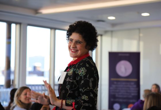 Smiling woman in a black and red outfit gestures while speaking to an audience.