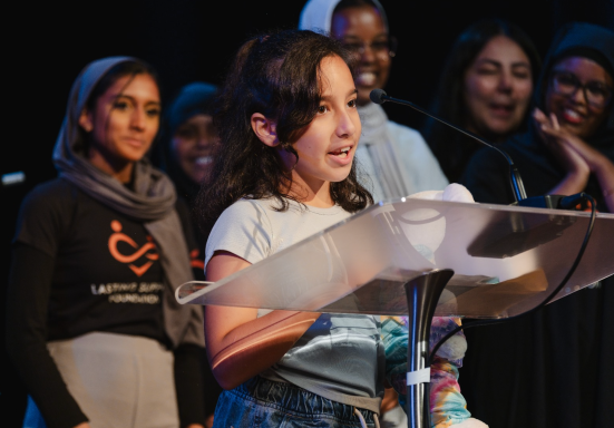 A young girl speaks at a podium, accompanied by smiling women in the background.