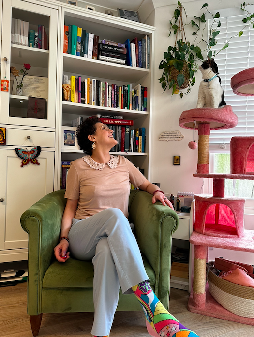 Person sitting in a green chair, looking up at a cat on a pink cat tree by a bookshelf.