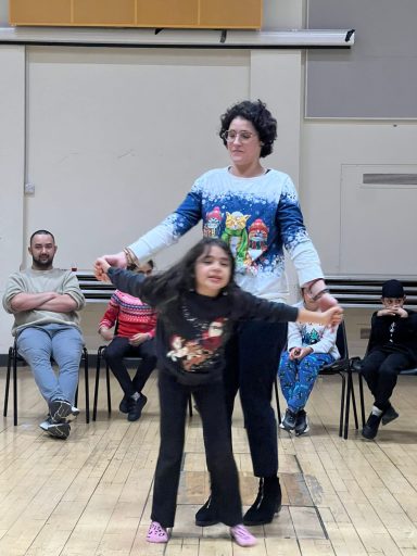 A woman in a festive sweater stands with a girl, both smiling joyfully, in a classroom setting.